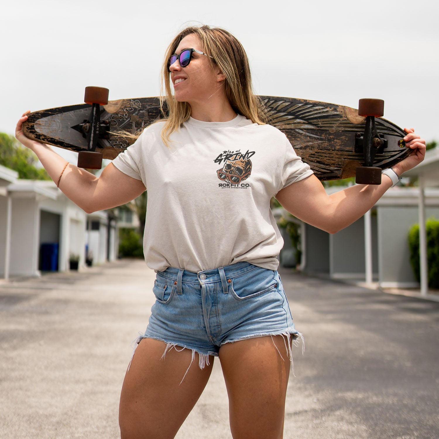 Female model outdoors wearing sand color Active Tee with 'Rise and Grind' graphic