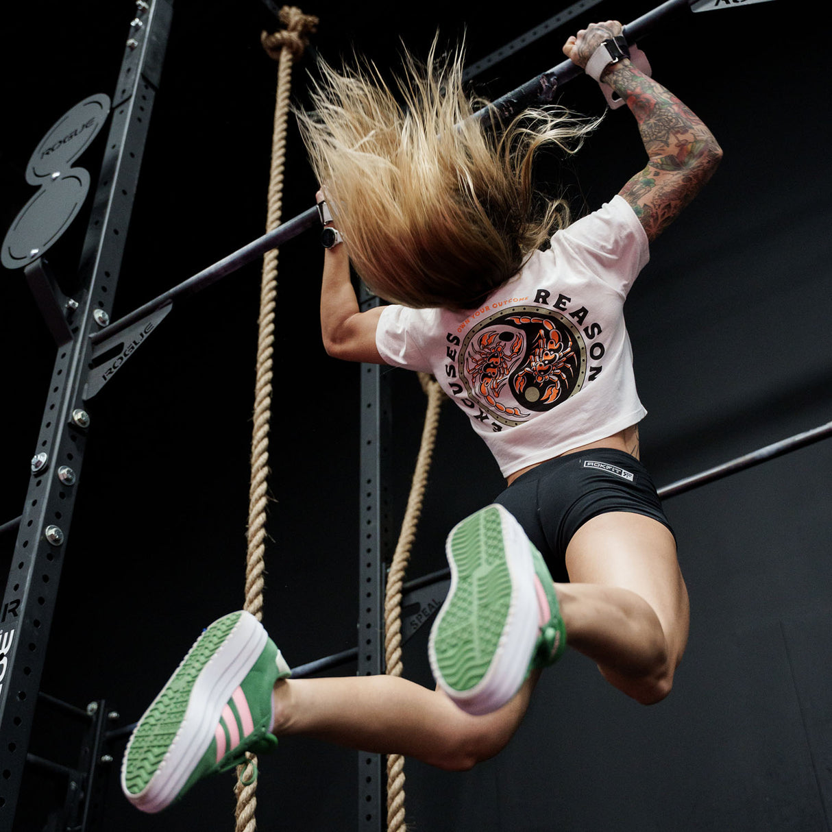 Female model doing a pull-up in a gym wearing white crop top with 'Excuses or Reasons' graphic