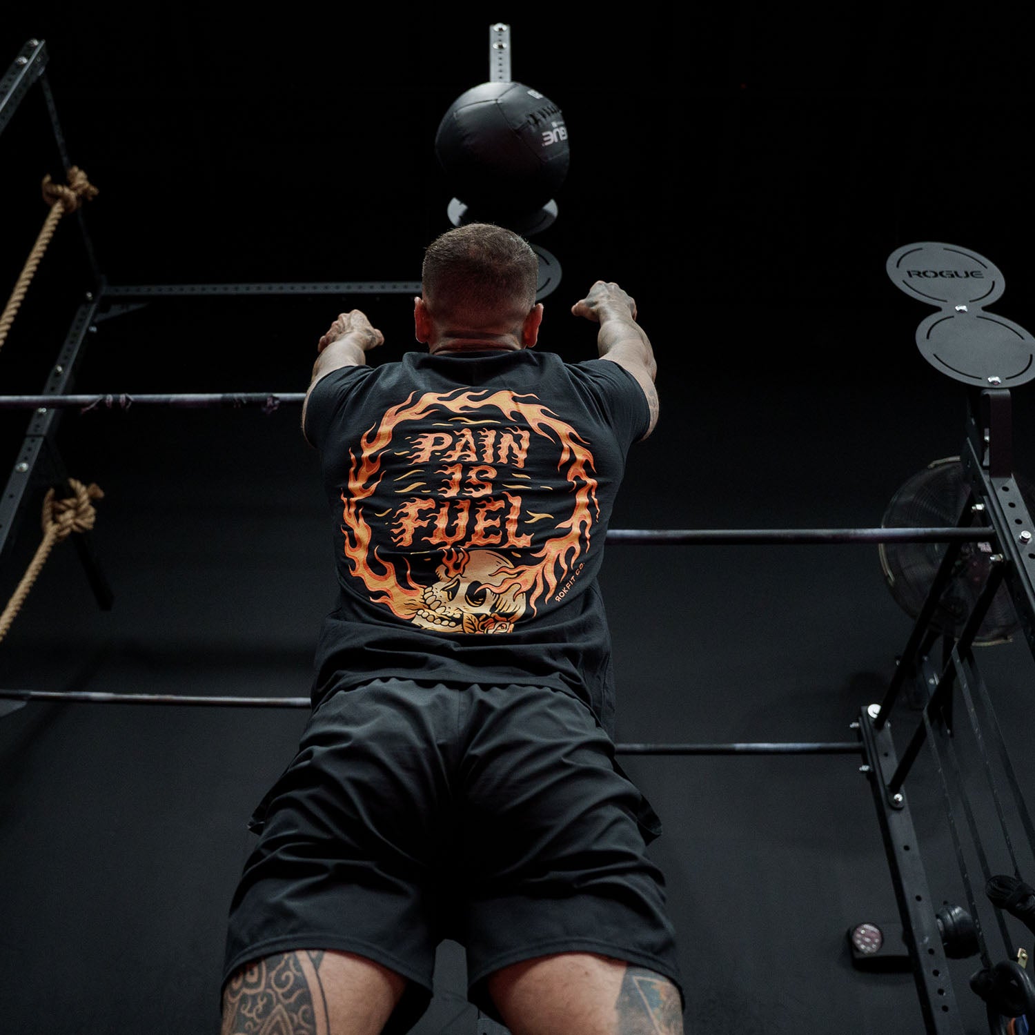 Male model doing a wall ball throw in a gym wearing Active Tee with 'Pain Is Fuel' graphic
