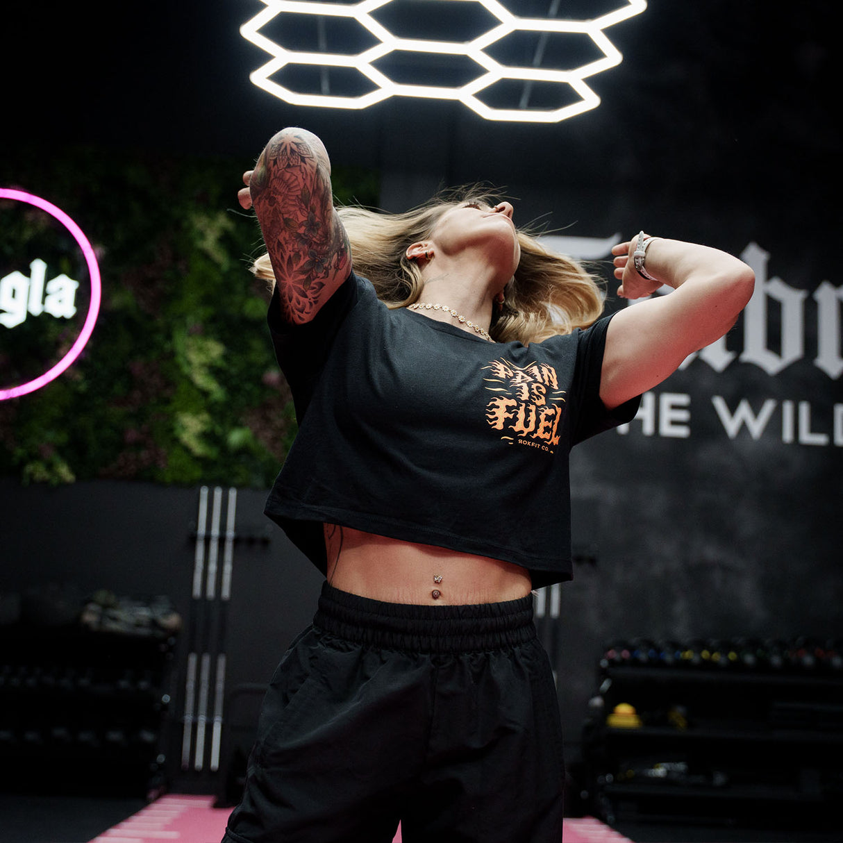 Female model in a gym wearing black crop top with 'Pain Is Fuel' graphic