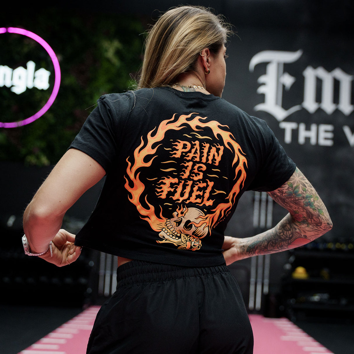 Female model in a gym wearing black crop top with 'Pain Is Fuel' graphic