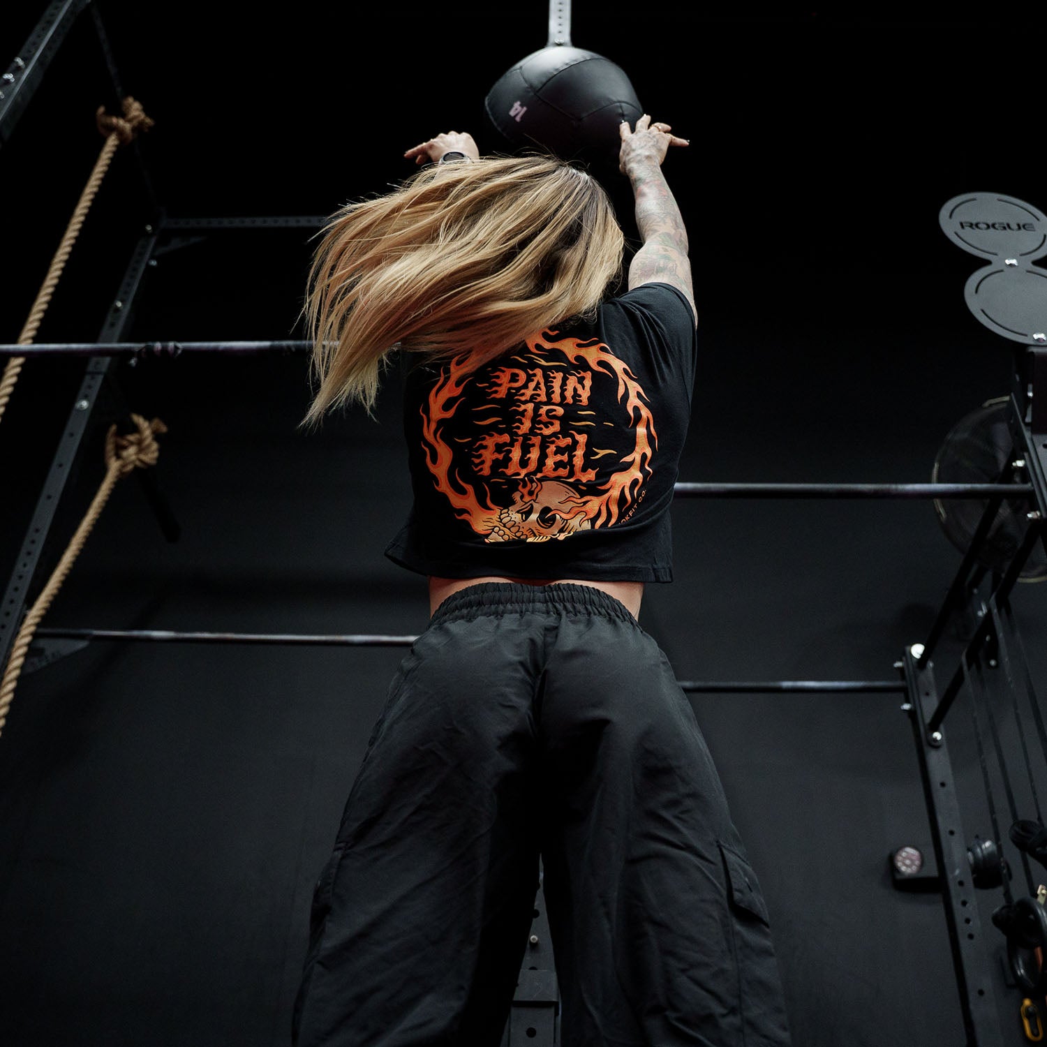 Female model doing a wall ball throw in a gym wearing black crop top with 'Pain Is Fuel' graphic